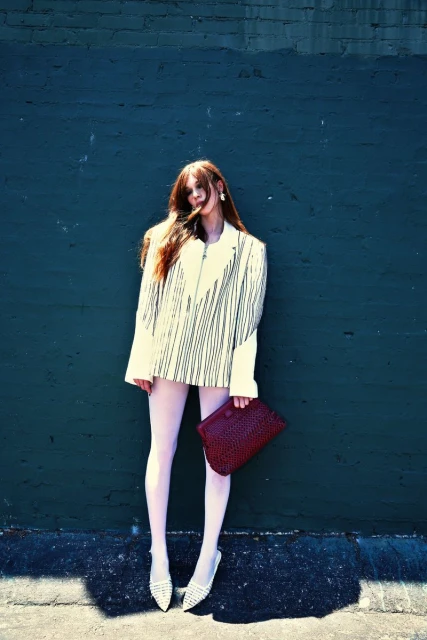 A stylish woman poses against a dark blue wall, wearing a striped blazer, light pink tights, and woven flats, holding a dark red woven clutch.