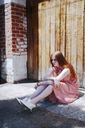 Red-haired model in a pink dress and white mules sitting outdoors against a wooden barn door and brick wall