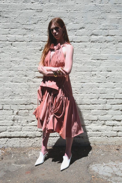 Elegant woman with red hair poses in a pink dress and white mule shoes against a white brick wall in an outdoor setting on a sunny day.