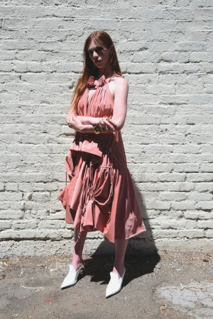 Elegant woman with red hair poses in a pink dress and white mule shoes against a white brick wall in an outdoor setting on a sunny day.