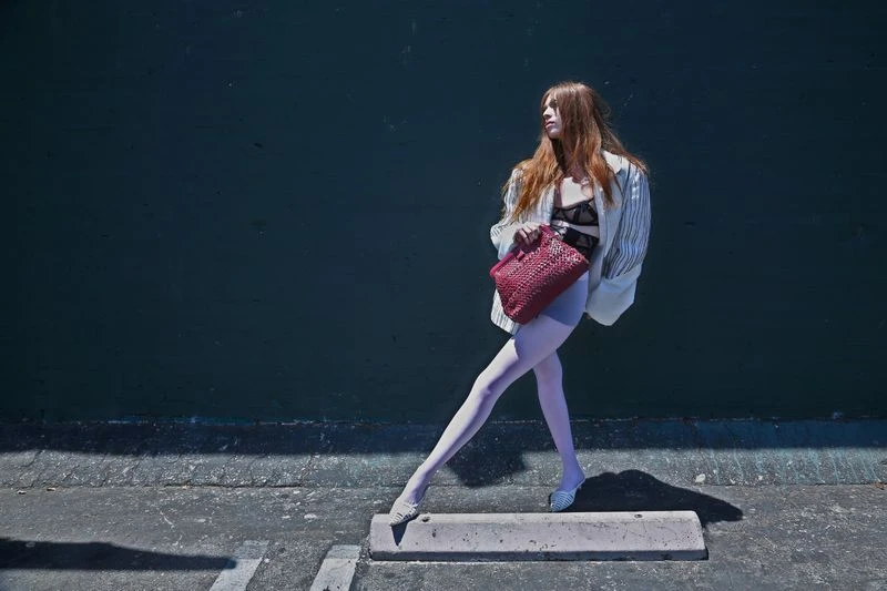 A young woman poses in front of a dark wall wearing a blazer, shorts, and tights, holding a red woven purse outdoors in sunlight.