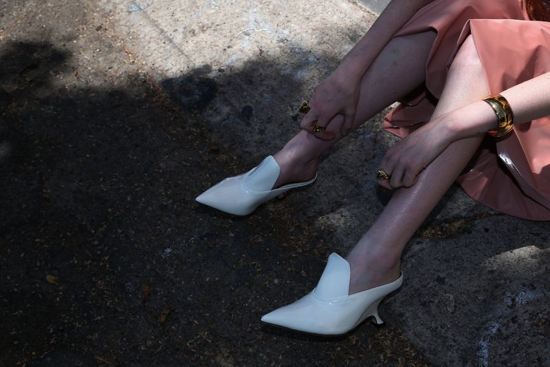 Elegant white heeled mules on a woman sitting outdoors in natural light, showcasing fashion footwear and accessories, emphasizing a sleek and modern style.