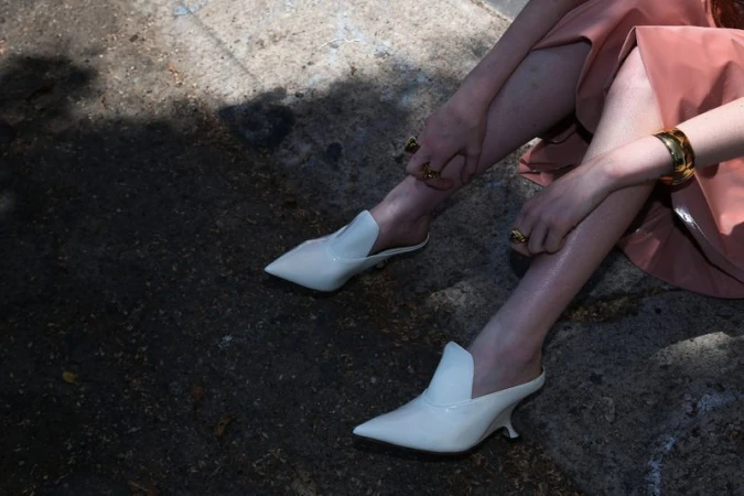 Elegant white heeled mules on a woman sitting outdoors in natural light, showcasing fashion footwear and accessories, emphasizing a sleek and modern style.
