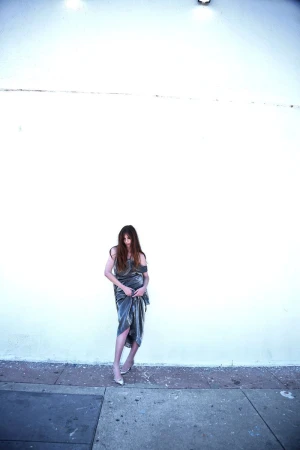 Woman in a metallic silver draped dress and heels standing against a large white wall on a sidewalk.