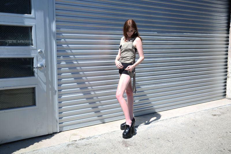 Adult woman in olive tank top and black shorts posing outside a gray metal garage door, wearing platform loafers, in full sunlight.
