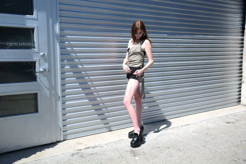 Adult woman in olive tank top and black shorts posing outside a gray metal garage door, wearing platform loafers, in full sunlight.