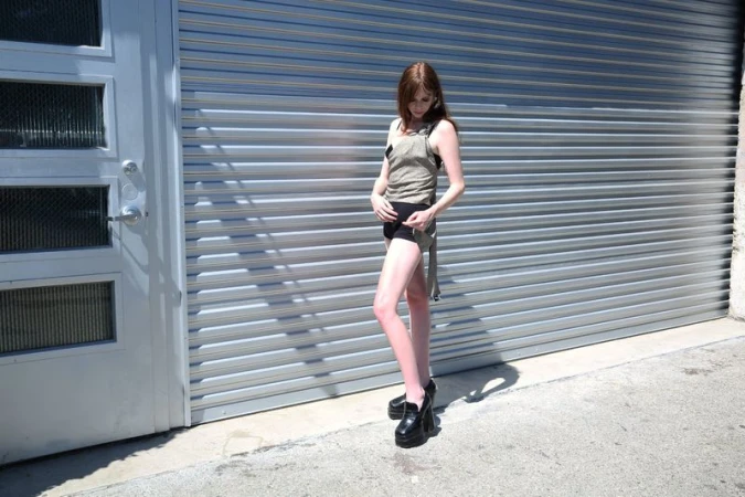 Adult woman in olive tank top and black shorts posing outside a gray metal garage door, wearing platform loafers, in full sunlight.