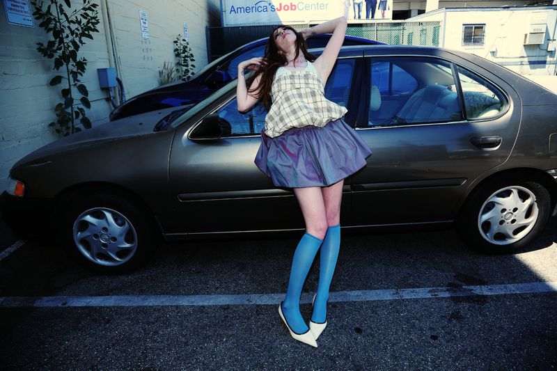 A young woman in a plaid top and blue skirt strikes a pose in a parking lot next to an older model car with a job center sign.