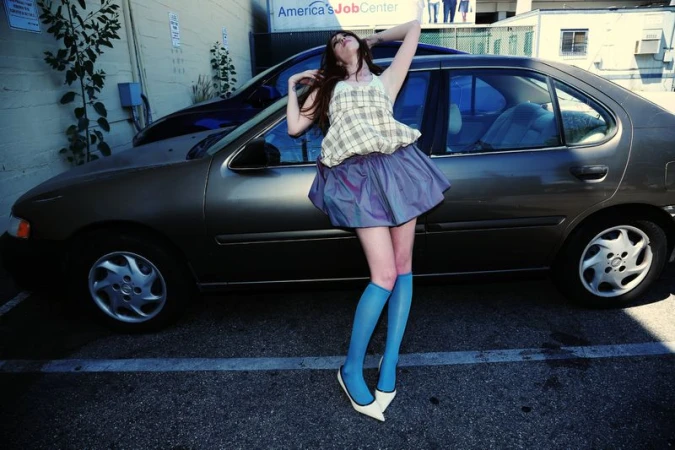 A young woman in a plaid top and blue skirt strikes a pose in a parking lot next to an older model car with a job center sign.