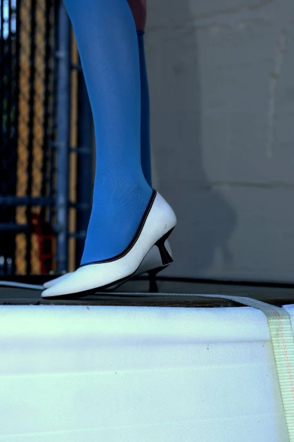 Close-up shot of a woman's lower legs and feet wearing blue tights and white high heel shoes with black trim standing on a surface.