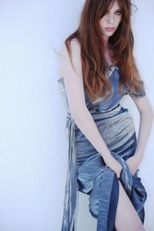 Woman with long red hair wearing a shimmering blue dress against a plain white wall