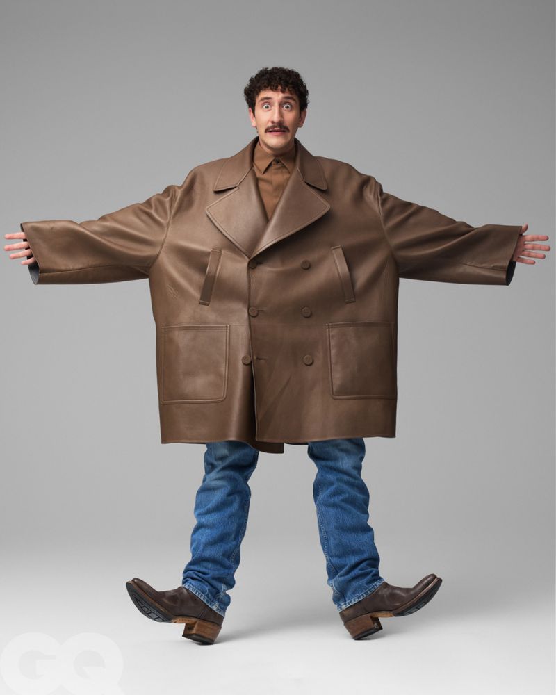 Full-length studio shot of an adult man in a brown leather double-breasted coat, blue jeans, and brown cowboy boots posing with arms outstretched.
