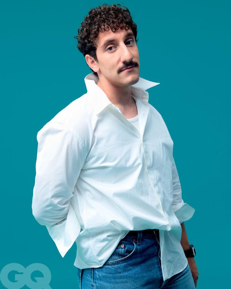 Portrait of a curly-haired adult male in a white shirt and blue jeans against a solid teal backdrop, vintage style, studio lighting.