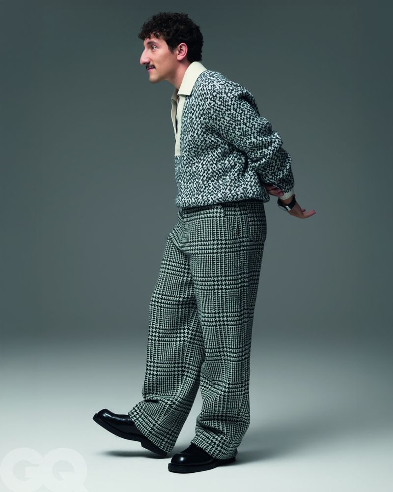 Man in patterned knit shirt and plaid pants strikes a pose in a studio. He has dark curly hair, a mustache, and black dress shoes.