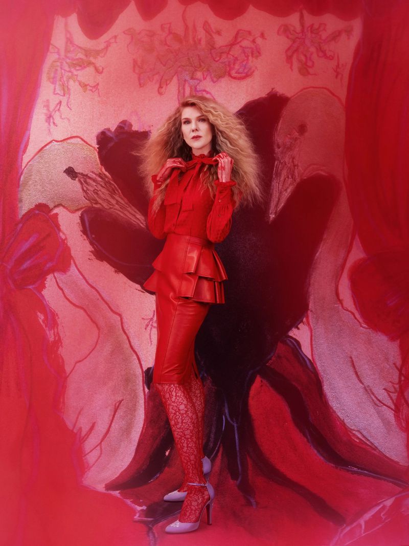 A fair-skinned adult female model in a red outfit stands in front of a red background with art. She is wearing high heels.