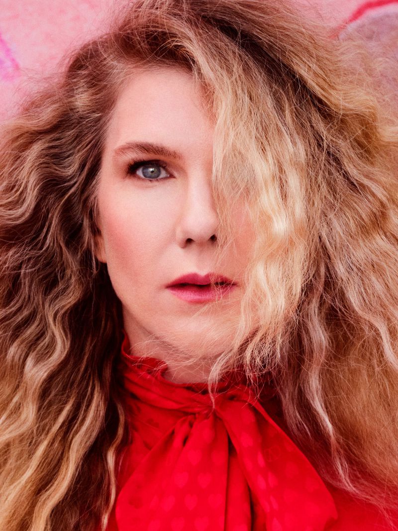 Portrait of an adult female with long blonde hair, wearing a red blouse with a tied neckline, set against a pink floral background.