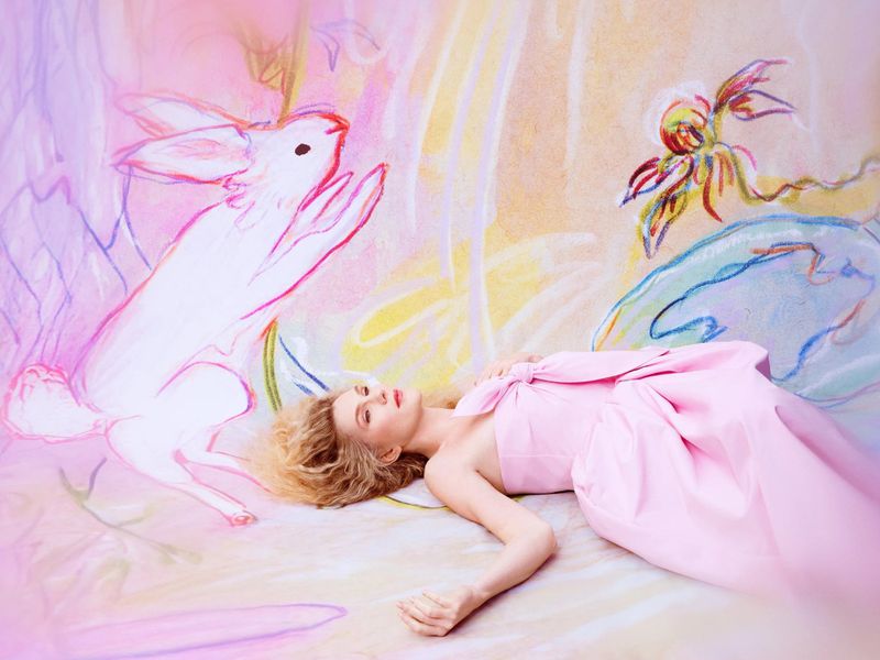 A woman in a pink dress poses lying down next to a white rabbit art piece, creating a whimsical and dreamy fashion editorial.