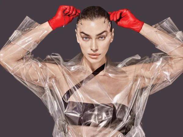 Model Irina Shayk wearing red leather gloves and a clear plastic coat against a dark grey background