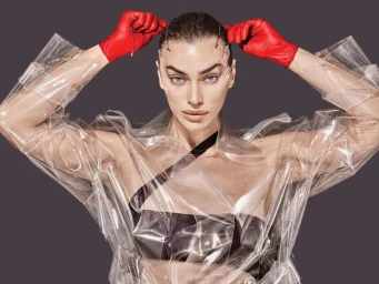Model Irina Shayk wearing red leather gloves and a clear plastic coat against a dark grey background