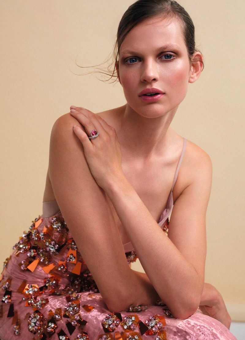 Portrait of a young adult female model in a light pink dress with decorative beading, wearing a pink gemstone ring, neutral background.