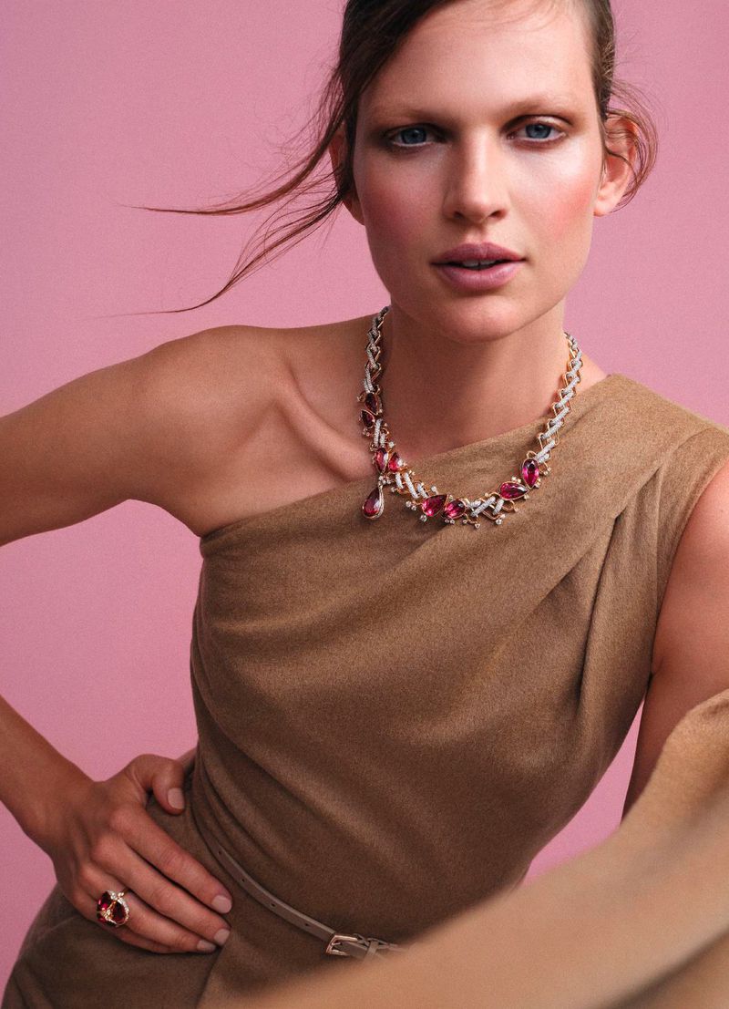 Elegant woman poses in a one-shoulder dress with a gemstone necklace and ring against a pink backdrop in a beauty fashion shot.
