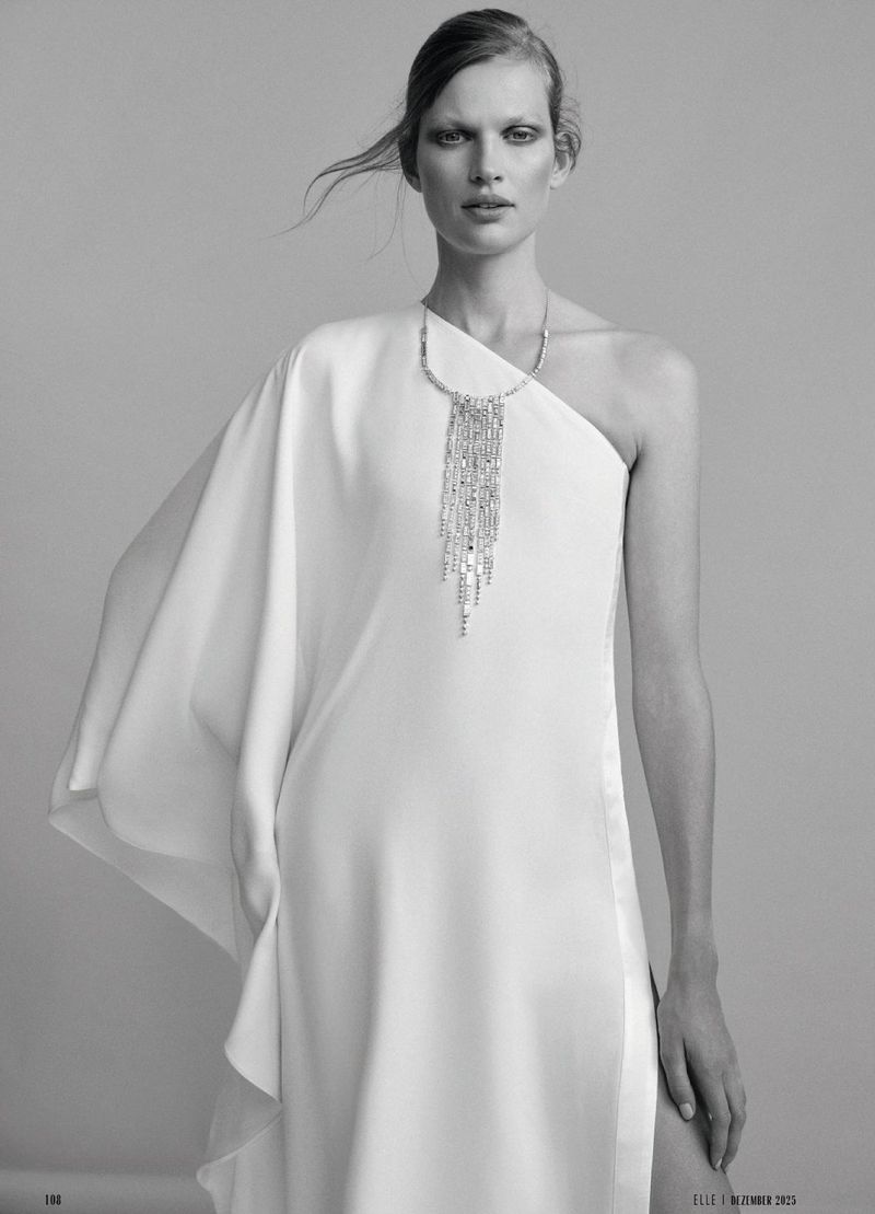 A woman with pale skin poses in a white dress and jeweled necklace in a studio. The black and white image has fashion and editorial style.