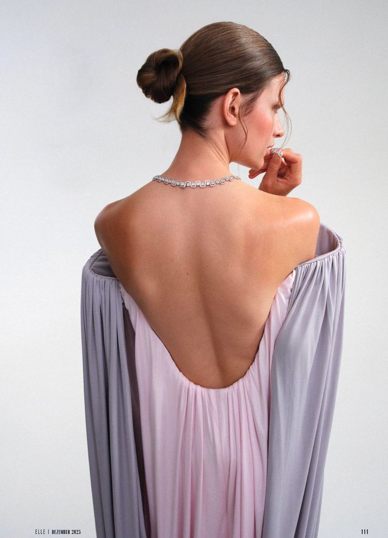 Back view of a female model in a lavender gown with a necklace, posing in a photo studio setting against a white backdrop.