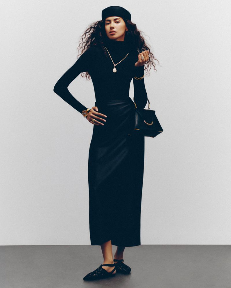 A woman wearing a black turtleneck, wrap skirt, and beret poses with a black handbag in a studio setting.