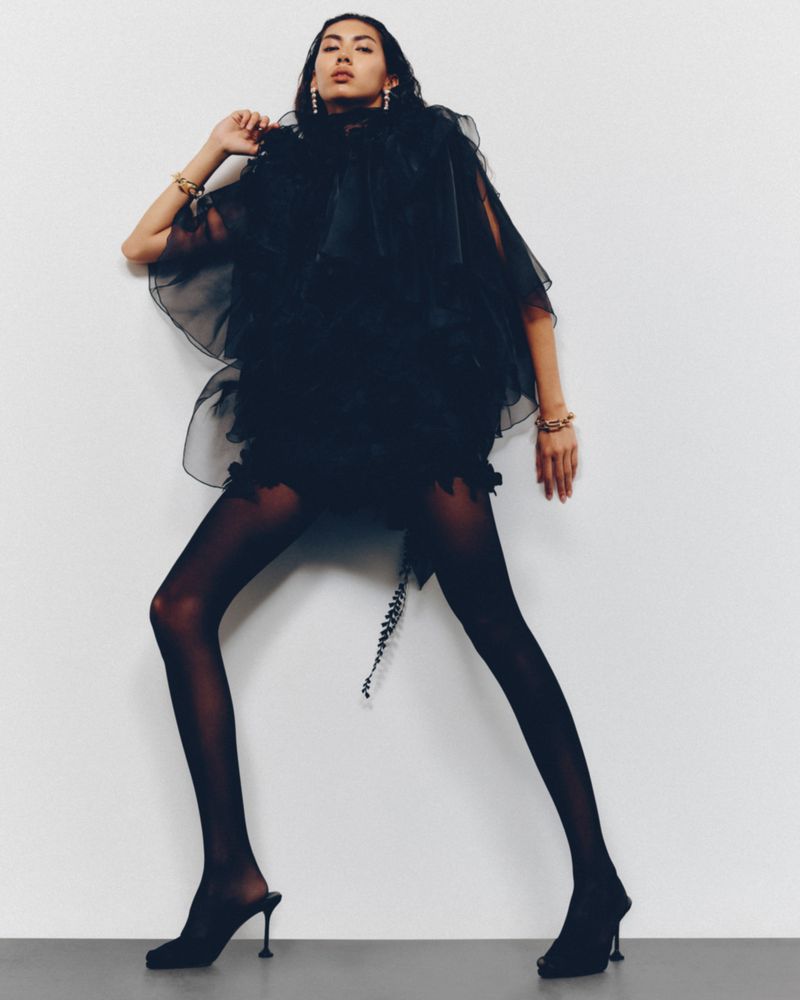 Elegant woman poses in a tiered black dress, black stockings, and black high heel mules against a clean white background for a fashion editorial.