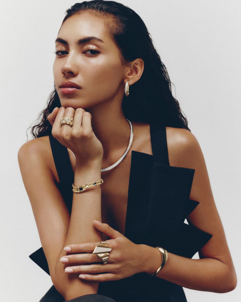 Portrait of a woman wearing an elegant black top and assorted gold and diamond jewelry against a neutral background.