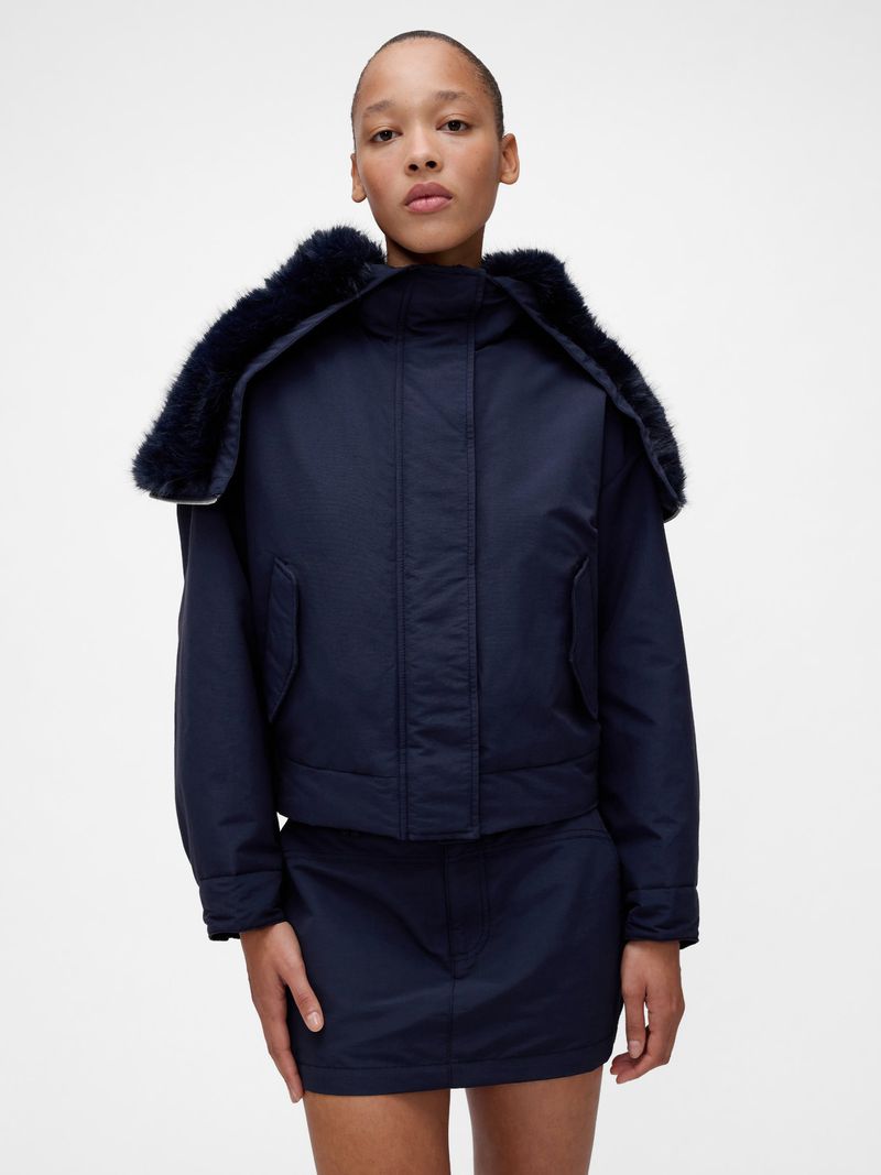 Woman in a navy blue jacket with a faux fur collar and a matching skirt poses against a white background in a studio setting.