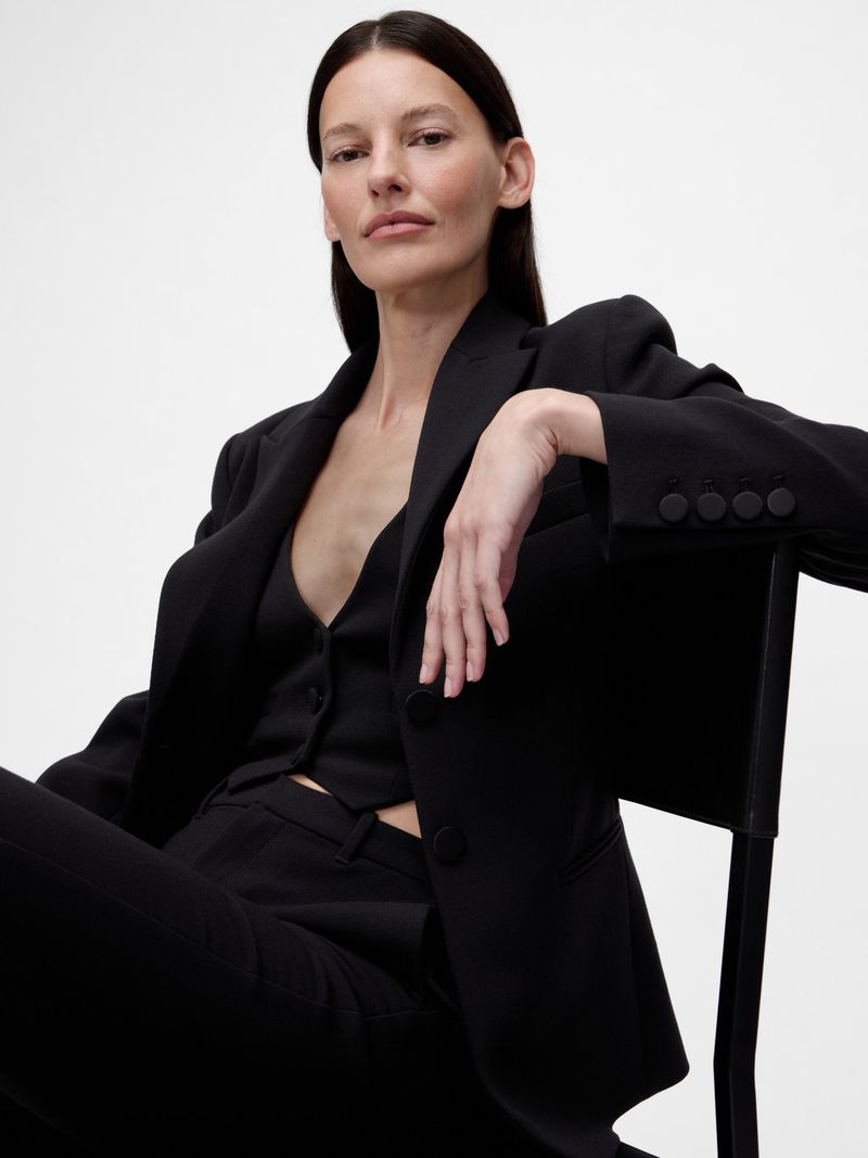 Model wearing a black tailored blazer and vest seated on a chair against a white background