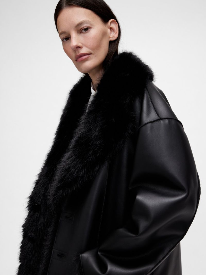 Adult woman posing in a black faux leather coat with a faux fur collar against a white studio backdrop in a fashion editorial-style photo.