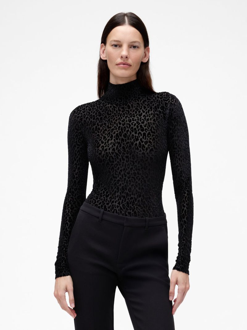 A fashionable adult female poses in a black turtleneck bodysuit with a leopard print design and black pants against a white backdrop.