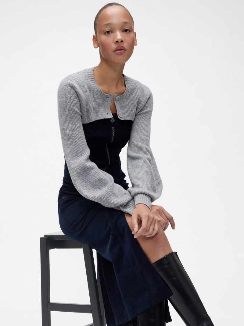 Elegant Black Woman in gray crop cardigan and navy dress sitting on black stool in a white studio with knee high boots.