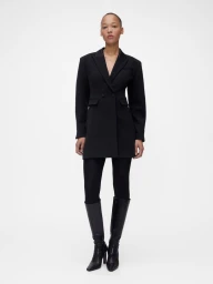 Model wearing a black tailored blazer coat and tall black boots against a plain white seamless studio background.