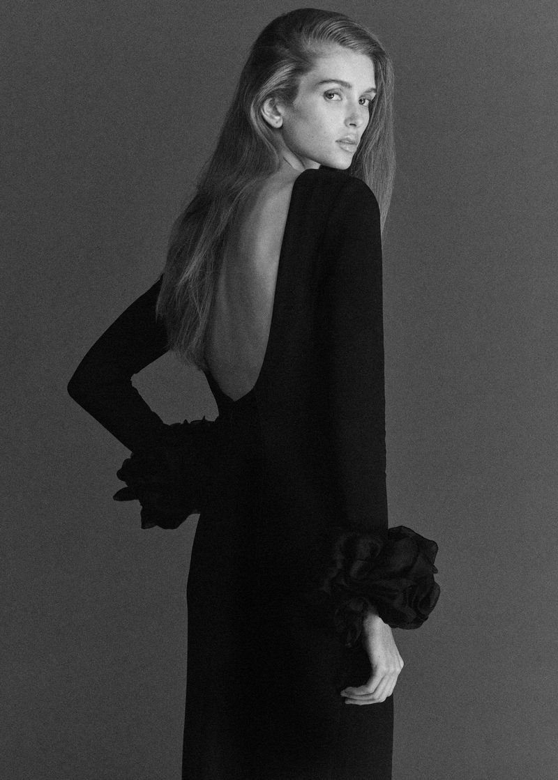 Monochrome studio portrait of a slender, light-skinned woman with long blond hair wearing a long-sleeved black open-back dress.