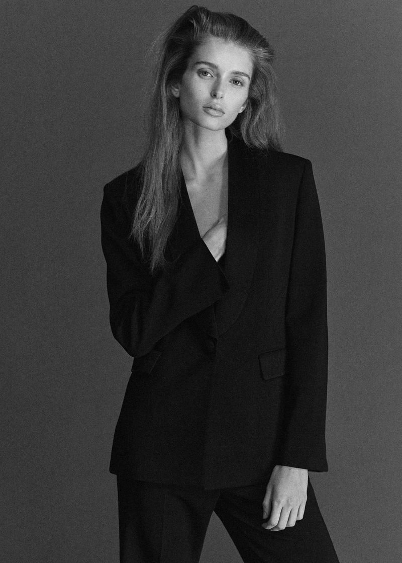 Striking black and white portrait of a fair-skinned woman in a stylish black blazer and pants, posing against a solid background.