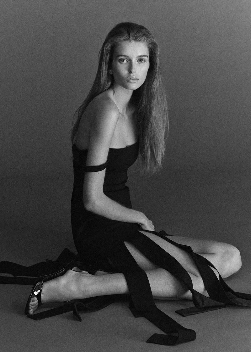 Model seated on a studio floor wearing a black dress with long ribbon ties, captured in monochrome.