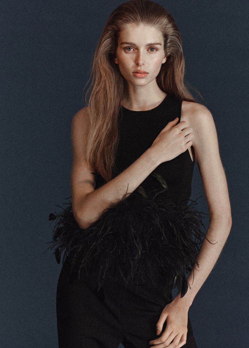 Portrait of a female fashion model in a black top with feather detail and black pants against a dark blue background in a studio.