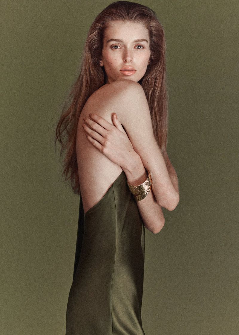 Portrait of a model with long brown hair, wearing a satin dress and gold bracelet, posing against an olive-green background in studio lighting.