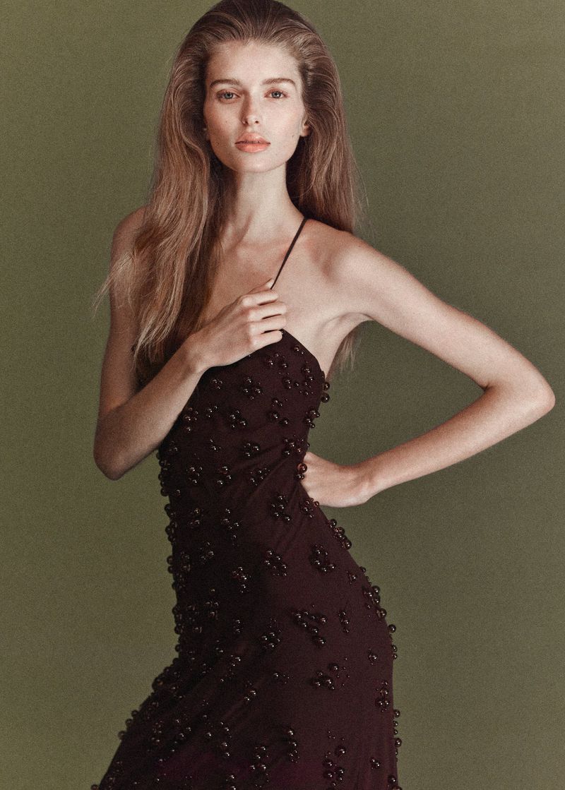 Elegant adult woman in a dark brown beaded evening gown posing against a muted green background in a fashion style photograph.
