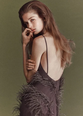 Model wearing a dark dress with feather trim posing against an olive green backdrop for Mango.