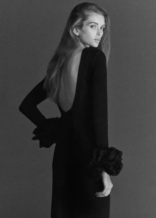 Model in a long black dress with voluminous ruffled cuffs looking over her shoulder against a plain grey background
