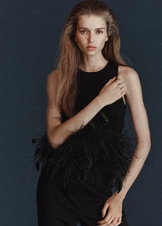 Model Stella Hanan wearing a black sleeveless top with feather trim against a dark backdrop