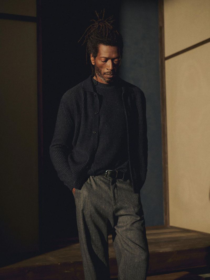 Adult male with dreadlocks poses indoors in dark buttoned sweater, grey trousers with black belt; indoor fashion editorial photography.