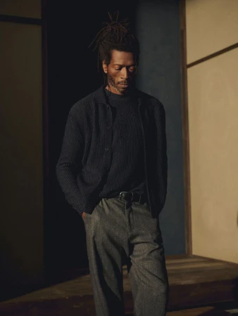 Adult male with dreadlocks poses indoors in dark buttoned sweater, grey trousers with black belt; indoor fashion editorial photography.
