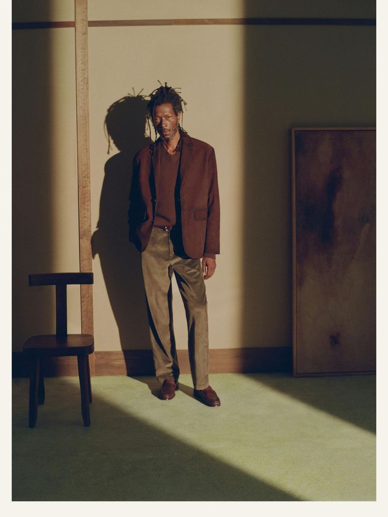 A man in a brown jacket and olive-green trousers poses in a room with a chair and artwork, lit by window light creating shadows.