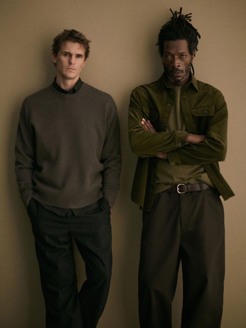 Two adult men wearing casual outfits pose against a neutral wall. One wears a brown sweater and the other a green corduroy jacket.