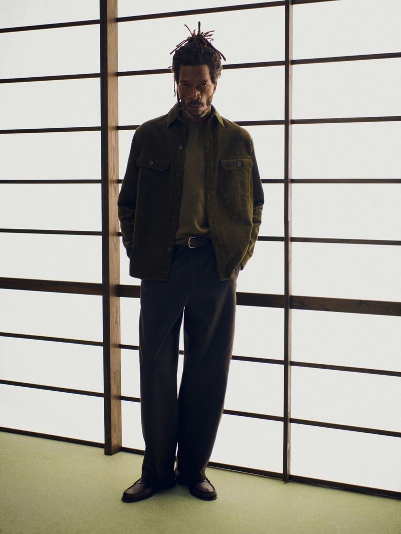 A man in an olive jacket and dark pants stands in front of a grid-patterned window, delivering a high-fashion portrait with neutral tones.
